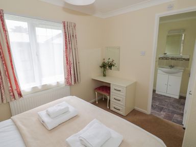 A bedroom with a bed with folded towels a desk with drawers a stool and a vase with flowers next to a window and a door leading to a bathroom at Daffodil in Eardisland