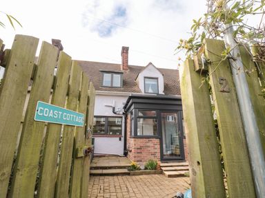 A wooden gate with Coast Cottage sign opening to a brick paved entrance and a house with brick and white walls at Coast Cottage in Bamburgh
