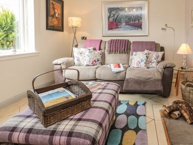 A living room with a sofa floral cushions and throw blankets a plaid ottoman with a wicker basket magazines a floor lamp a framed picture a colorful rug and a pile of firewood at Coast Cottage in Bamburgh