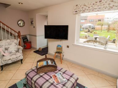 A living room with armchair a television on the wall and a large window showing a garden at Coast Cottage in Bamburgh