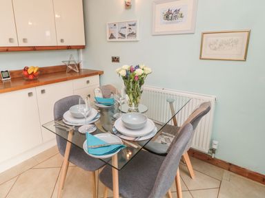 A dining area with a glass table set with plates bowls and glasses with chairs around and a kitchen counter with fruit bowl at Coast Cottage Bamburgh