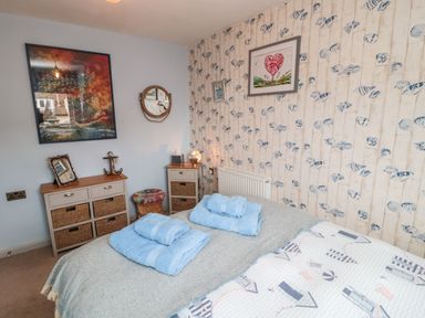 A bedroom with a bed, blue towels, two dressers with baskets, framed pictures, and wallpaper with shells at Coast Cottage in Bamburgh
