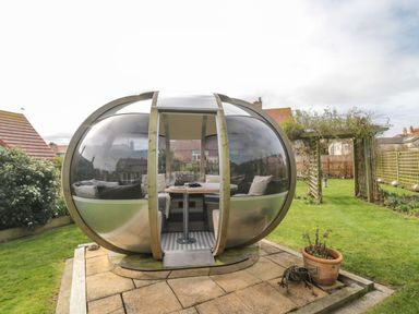 A transparent spherical garden pod with cushions and a table inside in a backyard with grass and a wooden pergola at Coast Cottage in Bamburgh