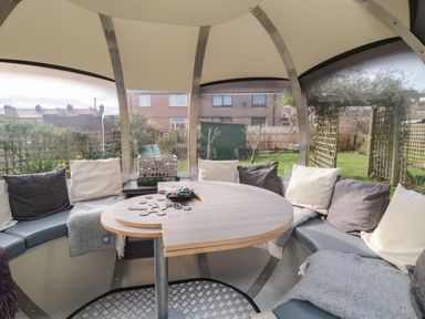 A round seating area with cushions and a table inside a gazebo overlooking a garden at Coast Cottage in Bamburgh