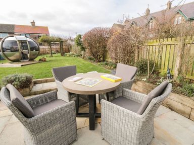 A garden with a round table and four chairs on a patio and a pod structure in the grass at Coast Cottage in Bamburgh