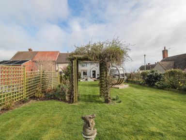 A garden with a wooden arch covered in vines a lawn sculpture a lawn with flower beds and a house in the background at Coast Cottage in Bamburgh