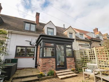 A small brick and glass conservatory attached to the back of terraced houses with a paved patio and wooden garden furniture at Coast Cottage in Bamburgh