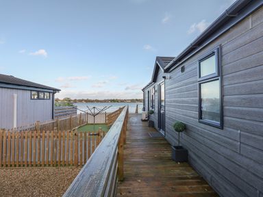 A wooden walkway beside a grey house with potted plants and a fenced garden with drying rack overlooking a lake at Lake View Lodge in Tallington