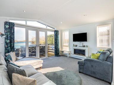 A living room with sofas a wall-mounted TV a fireplace and large glass doors overlooking water at Lake View Lodge in Tallington