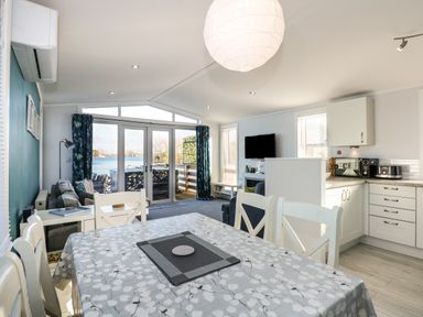 A dining area with a table and chairs next to a kitchen and living room with a sofa and television overlooking a lake at Lake View Lodge in Tallington