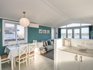 A dining area with table and chairs next to a living space with sofa and windows overlooking water at Lake View Lodge in Tallington