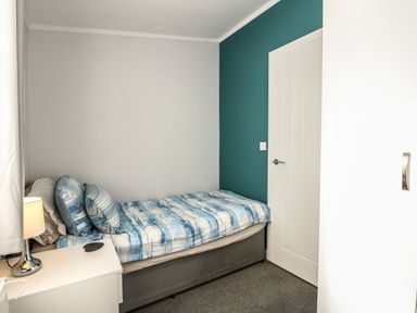 A small bedroom with a single bed blue and white bedding a white nightstand with a lamp a closed door and a teal accent wall at Lake View Lodge in Tallington