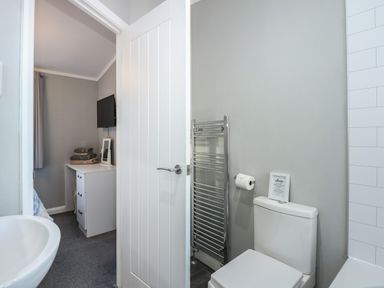 A bathroom with a toilet sink towel rack and an open door showing a bedroom with a desk and a wall-mounted TV at Lake View Lodge in Tallington