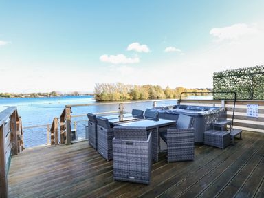 An outdoor wooden deck with a dining table set and a hot tub overlooking a river at Lake View Lodge in Tallington