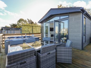 An outdoor deck with wicker chairs and glass table next to a hot tub and a cabin with open glass doors at Lake View Lodge in Tallington