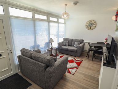 A living room with two grey sofas a small table with a lamp a patterned rug a dining table with chairs a wall clock and a television at 114 Cherry Park in Chapel St Leonards