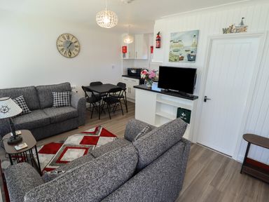A living room with grey sofas a dining table with four black chairs a TV on a white cabinet and a kitchen area at 114 Cherry Park in Chapel St Leonards