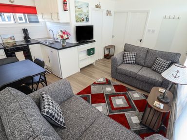 A living room with two gray sofas a patterned rug a small wooden side table with a lamp a black dining table with chairs and a kitchen area at 114 Cherry Park in Chapel St Leonards
