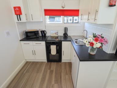 A kitchen with white cabinets black countertops a stove microwave kettle sink and flowers in a jug on the counter at 114 Cherry Park in Chapel St Leonards