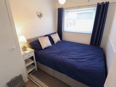 A bedroom with a bed covered in blue bedding and two pillows a bedside table with a lamp and decor a window with blue curtains and a wall hanging at 114 Cherry Park in Chapel St Leonards