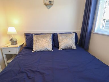 A bedroom with a bed with blue bedding and silver pillows a bedside table with a lamp and a window with blue curtains at 114 Cherry Park in Chapel St Leonards