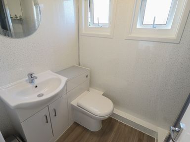 A bathroom with a sink vanity unit, toilet, two windows, and a round mirror on the wall at 114 Cherry Park in Chapel St Leonards