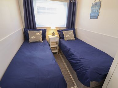 A bedroom with two single beds with blue bedding a white nightstand and a lamp at 114 Cherry Park in Chapel St Leonards