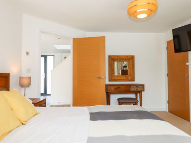 A bedroom with a bed yellow pillows bedside table lamp wooden desk mirror stool and wall mounted television at Pebble Ridge View in Westward Ho