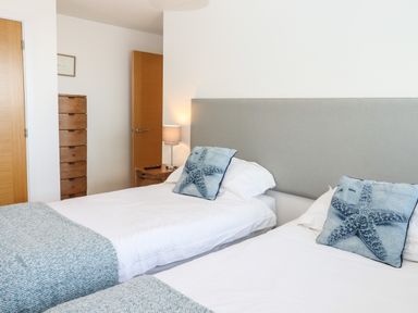 A bedroom with two single beds each with a starfish pillow and a gray headboard a wooden chest of drawers a wooden door and a bedside table with a lamp at Pebble Ridge View in Westward Ho
