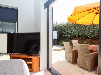 A room with a television on a wooden stand and an open door leading to a patio with wicker chairs and an orange umbrella at Pebble Ridge View in Westward Ho