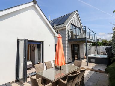 An outdoor patio with a glass dining table and wicker chairs an orange umbrella a hot tub a balcony and a white house at Pebble Ridge View in Westward Ho