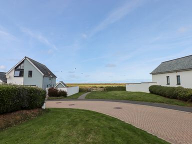 A paved pathway between two houses with grass and shrubs leading to a gate and open fields at Pebble Ridge View in Westward Ho