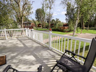 A wooden deck with white railing and black chairs overlooking grass and trees at 18 Woodlands Retreat in Tattershall
