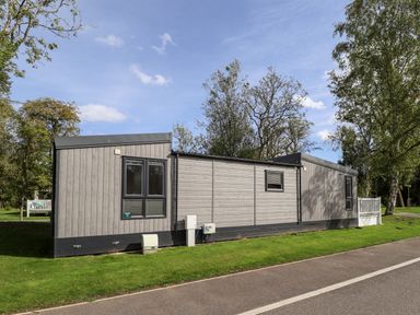 A single-story modular building with gray siding and multiple windows surrounded by grass and trees at 18 Woodlands Retreat in Tattershall