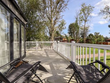 A wooden deck with two black lounge chairs next to glass doors overlooking trees and grass at 18 Woodlands Retreat in Tattershall
