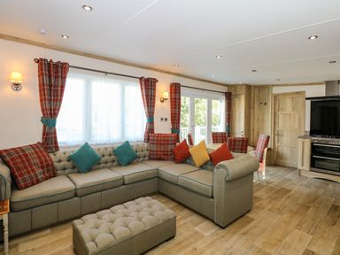 A living room with an L-shaped sofa cushions and plaid curtains near a kitchen with wooden cabinets at 18 Woodlands Retreat in Tattershall