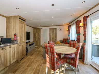 A dining area with a round table and four plaid chairs next to a living room with a sofa a television and wooden cabinets at 18 Woodlands Retreat in Tattershall
