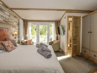 A bedroom with a bed and pillows towels on it two armchairs by a glass door a wall mounted TV and a wooden wardrobe at 18 Woodlands Retreat in Tattershall