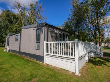 A grey mobile home with white railing on a green lawn with trees at 18 Woodlands Retreat in Tattershall