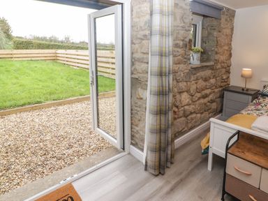 A bedroom with a bed a nightstand with a lamp a small chest of drawers a stone wall and an open glass door leading to a garden at The Nest in Amroth
