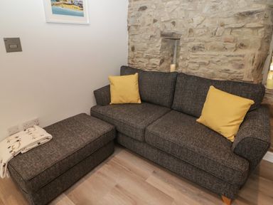 A gray sofa with two yellow cushions and a matching ottoman with a blanket in a room with a stone wall at The Nest in Amroth