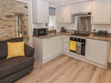 A kitchen with white cabinets a microwave a sink an oven and a toaster next to a gray sofa with a yellow cushion at The Nest in Amroth