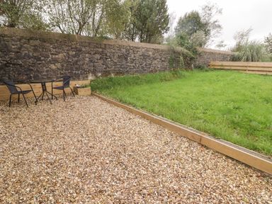 A small patio area with gravel floor a black metal table and two chairs next to a grass lawn and stone wall at The Nest in Amroth