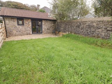A grassy garden area with stone walls and a small stone building with glass doors and a patio at The Nest in Amroth