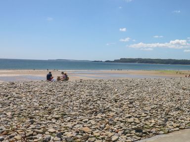 A rocky beach with people sitting and walking near the water and a distant landmass under a blue sky at The Nest in Amroth