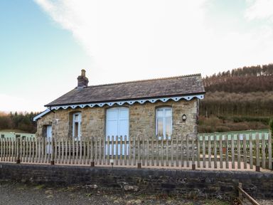 A stone cottage with white doors and windows behind a wooden picket fence with trees and hills in the background at Railway Station Cottage in New Radnor near Kington