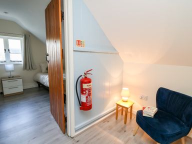 A corner with a blue chair a small wooden table with a lamp and a book a fire extinguisher mounted on the wall and a bedroom visible through an open wooden door at Railway Station Cottage in New Radnor near Kington