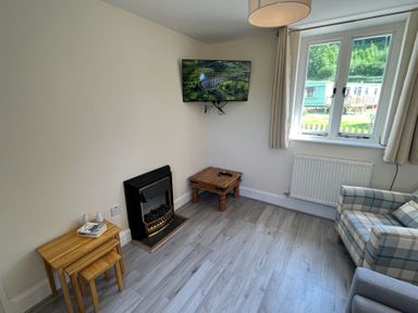 A small living room with a mounted television above a fireplace two nested wooden tables a wooden side table with remotes a plaid armchair next to a window with curtains at Railway Station Cottage in New Radnor near Kington