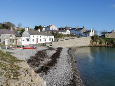 A pebble beach with boats near a stone seawall and houses in a coastal village at 3 Moranedd in Moelfre