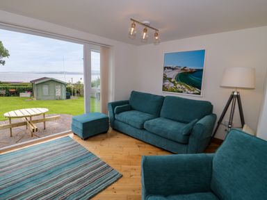 A living room with blue sofas and a rug next to a glass door overlooking a garden with a shed at 3 Moranedd in Moelfre